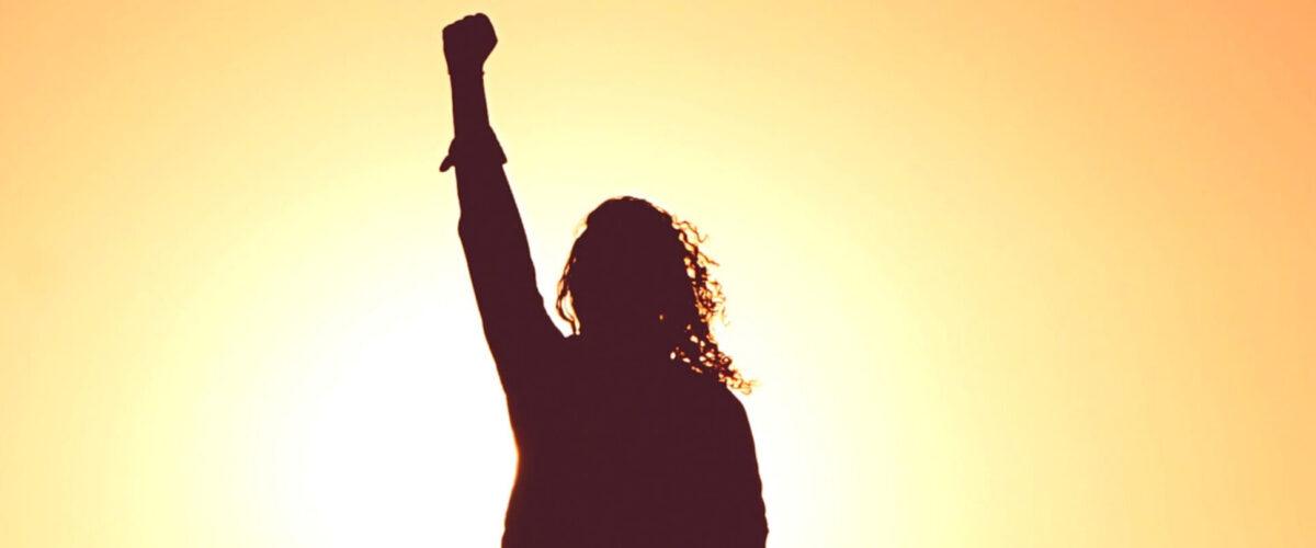 A silhouette of a woman against the sun with her arm up in the air in defiance.