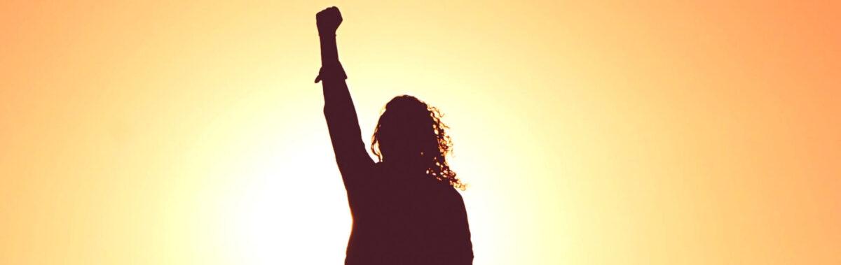 A silhouette of a woman against the sun with her arm up in the air in defiance.